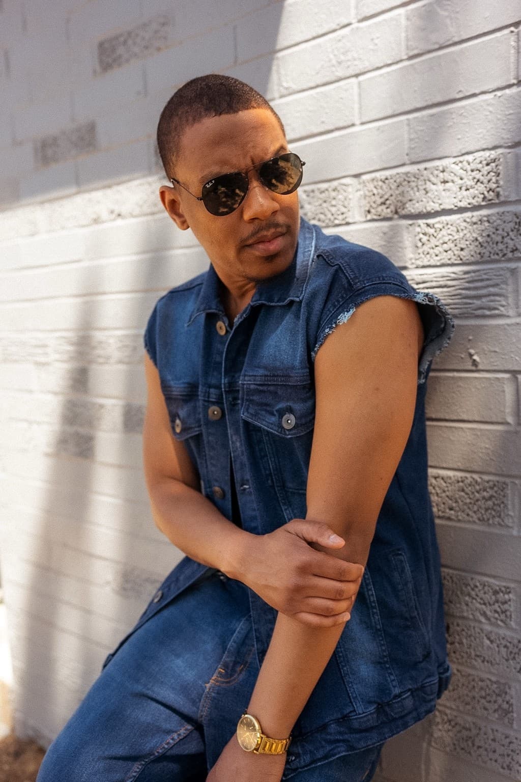Joseph Johnson (Speezy OTB), founder of International Playing Field, leaning confidently against a white brick wall wearing sunglasses and a denim vest